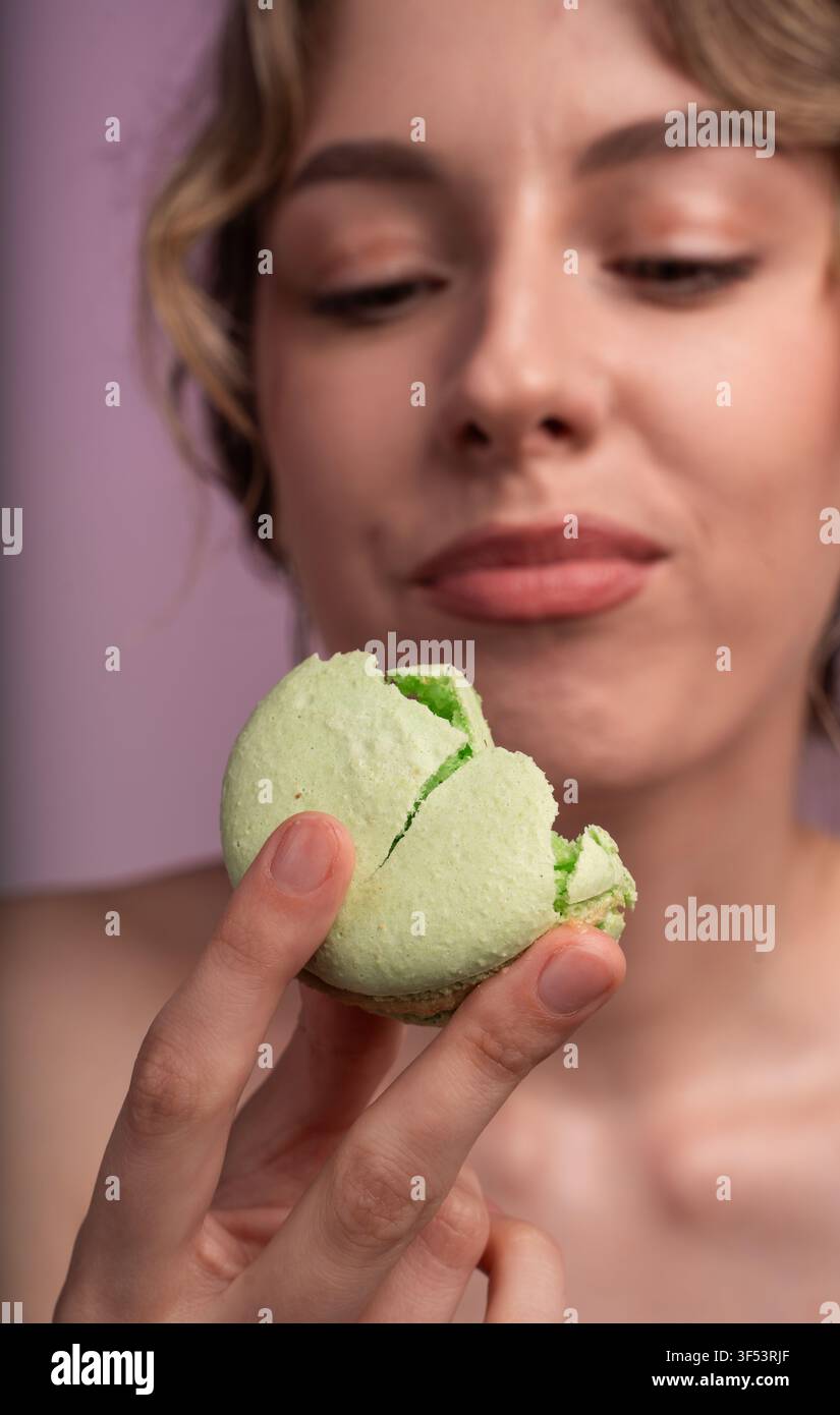 Giovane donna che si gode un momento dolce, tenendo e mangiando un macaron verde su uno sfondo lilla morbido Foto Stock