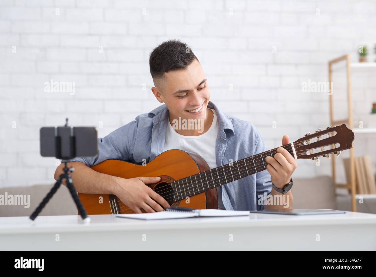 Giovane studente che impara a suonare la chitarra durante l'autoisolamento a casa o con un tutor online. Ragazzo sorridente guarda la chitarra e fa una lezione video per i suoi follower Foto Stock