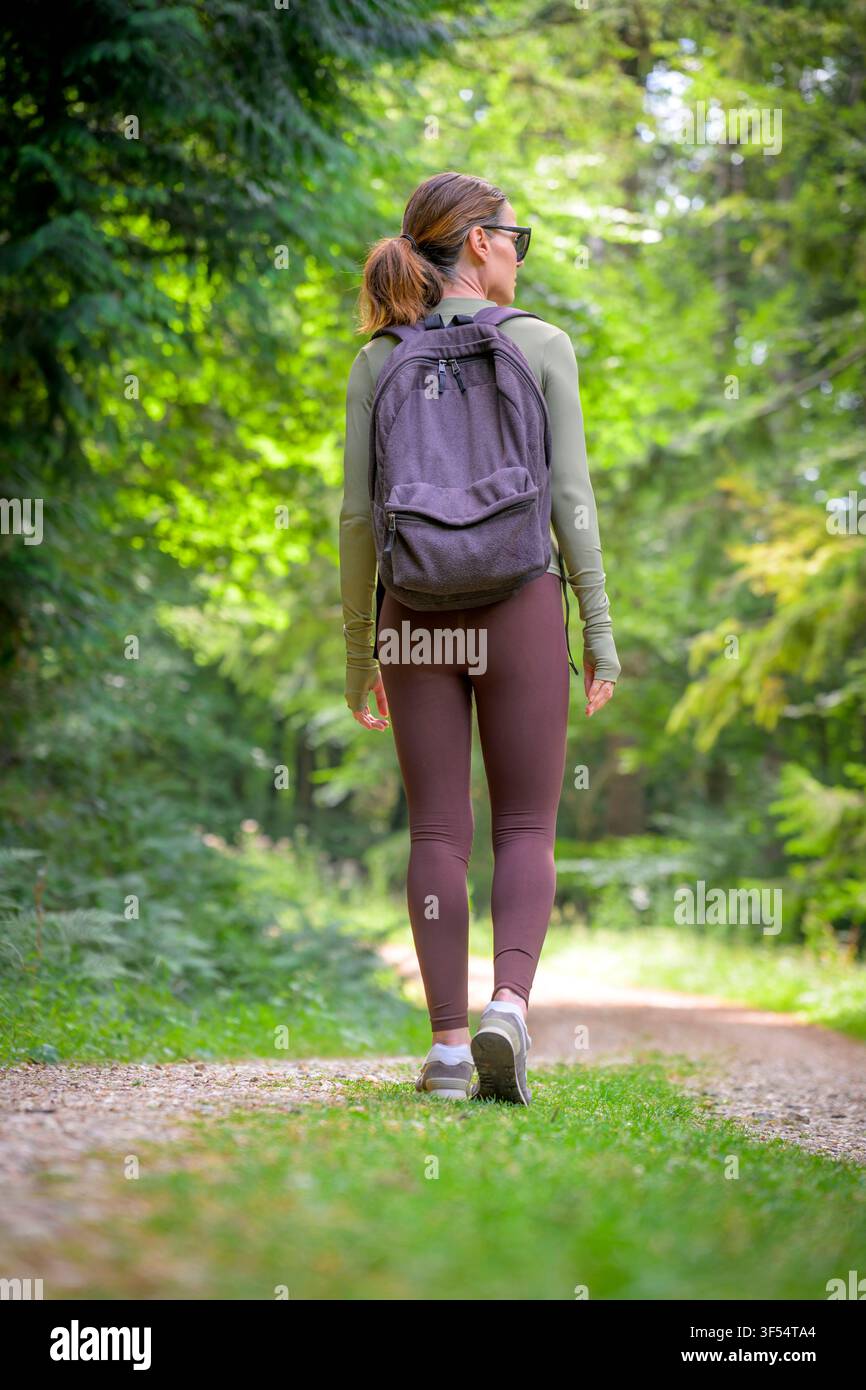 Escursionista femminile che cammina lungo un sentiero boschivo illuminato dal sole indossando uno zaino Foto Stock