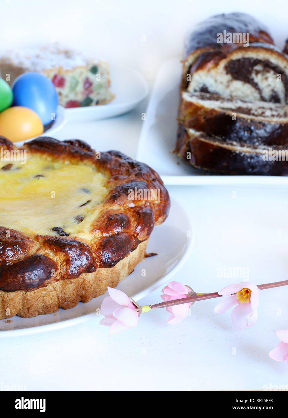 Tradizionale torta pasquale rumena Pasca con formaggio dolce e uvetta, accanto a uova colorate e fiori primaverili su tavolo bianco. Stagione pasquale e festiva Foto Stock