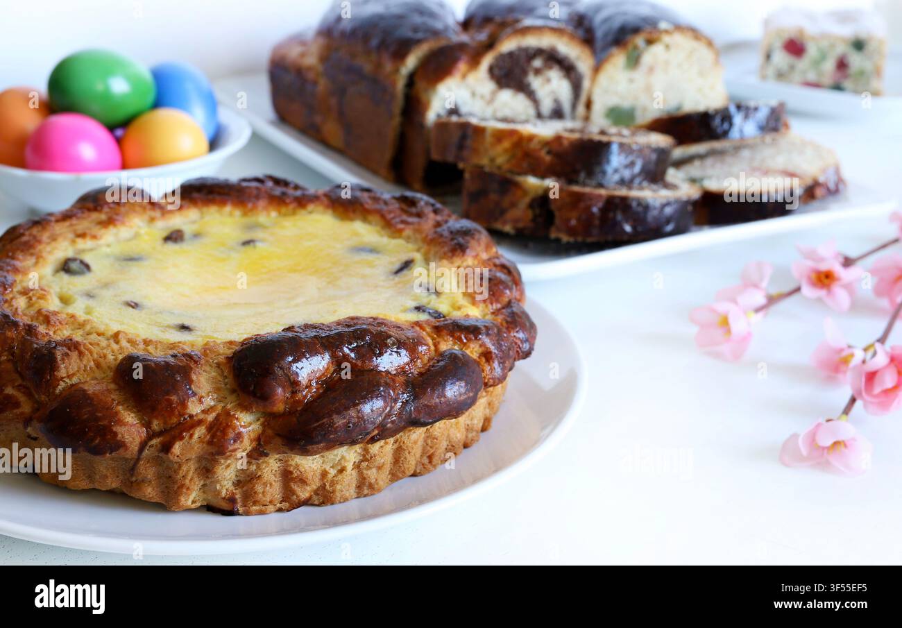 Tradizionale torta pasquale rumena Pasca con formaggio dolce e uvetta, accanto a uova colorate, fiori primaverili e pane dolce Cozonac su tavolo bianco. Foto Stock