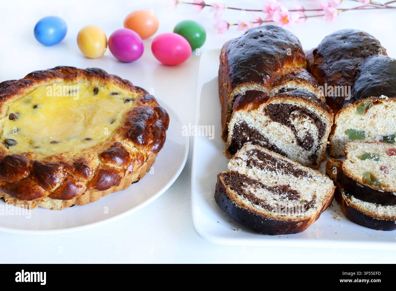 Tradizionale torta pasquale rumena Pasca con formaggio dolce, uvetta e pane dolce Cozonac, accanto a uova colorate e fiori primaverili su tavolo bianco. Foto Stock