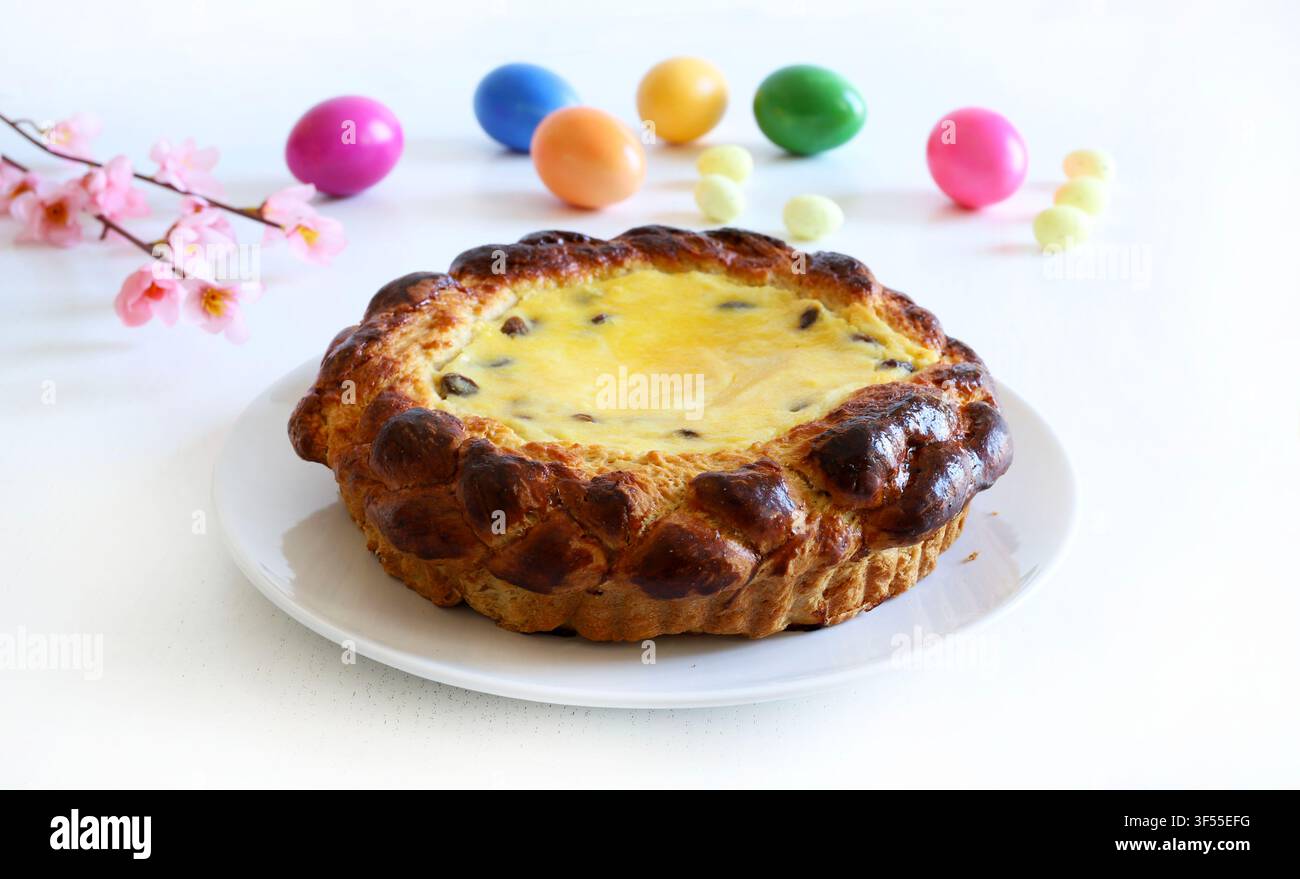 Tradizionale torta pasquale rumena Pasca con formaggio dolce e uvetta, accanto a uova colorate e fiori primaverili su tavolo bianco. Pasqua e stagione delle feste. Foto Stock