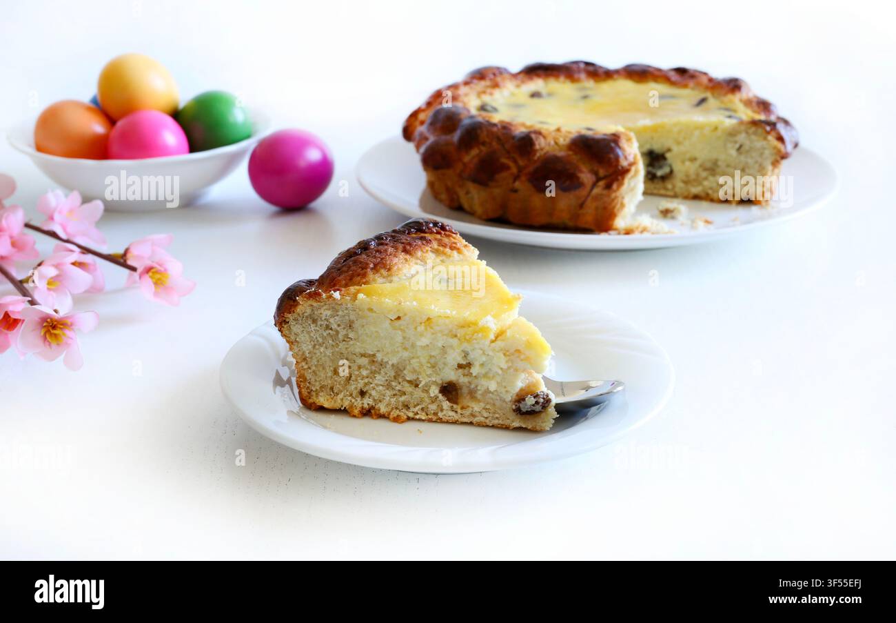 Fetta di tradizionale torta rumena Pasca con ricotta e uvetta, accanto a uova colorate su tavolo bianco. Concetto di Pasqua e festività. Foto Stock