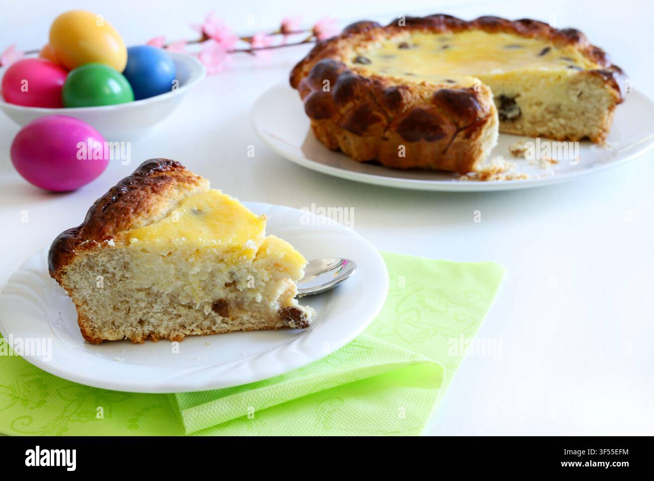 Fetta di tradizionale torta rumena Pasca con ricotta e uvetta, accanto a uova colorate su tavolo bianco. Concetto di Pasqua e festività. Foto Stock