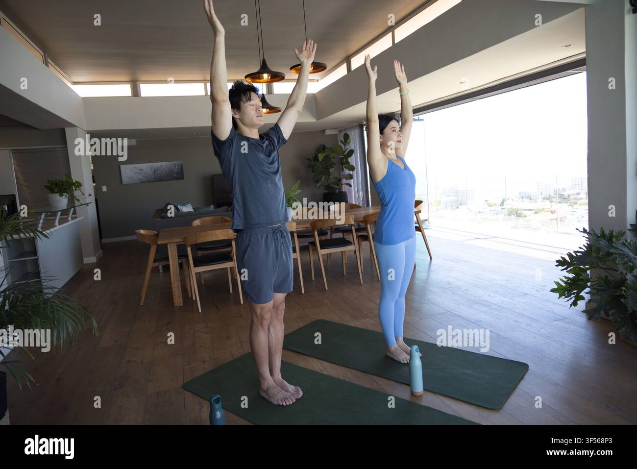 Coppia asiatica che pratica yoga in piedi in una casa moderna su materassini verdi con abbigliamento sportivo e bottiglie Foto Stock