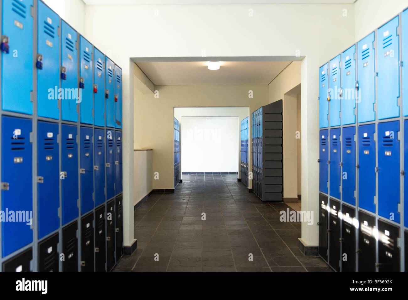 Corridoio libero con armadietti superiori blu, inferiori scuri, serrature, prese d'aria, corridoio piastrellato che conduce a una porta Foto Stock