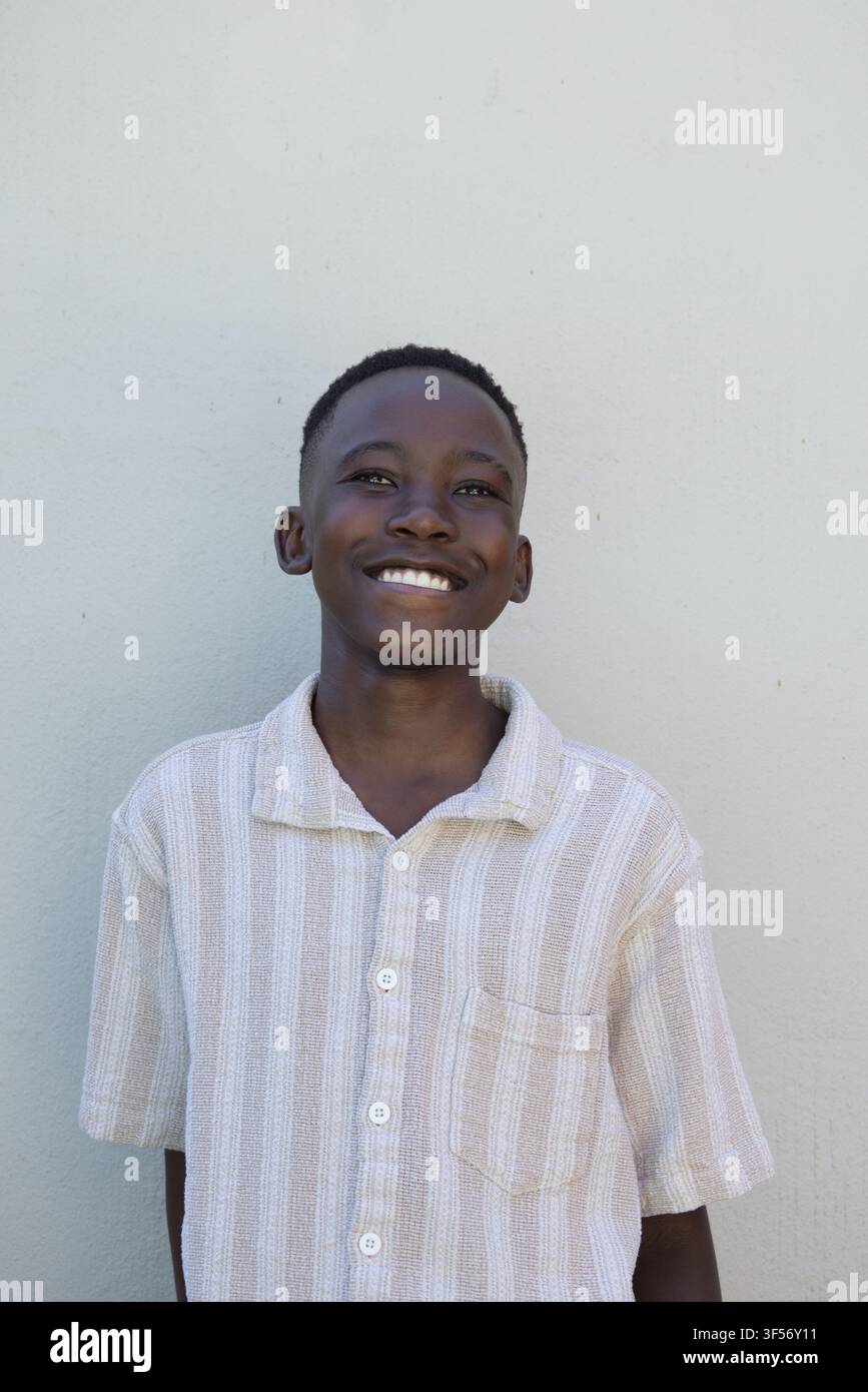 Ragazzino africano sorridente, indossa una camicia beige a maniche corte con tasca sul petto davanti a un muro grigio Foto Stock