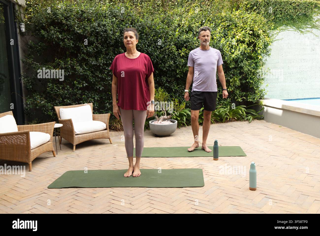 Coppia matura che pratica esercizi su materassini da yoga verdi, bottiglie d'acqua sul patio, indossa abbigliamento sportivo Foto Stock