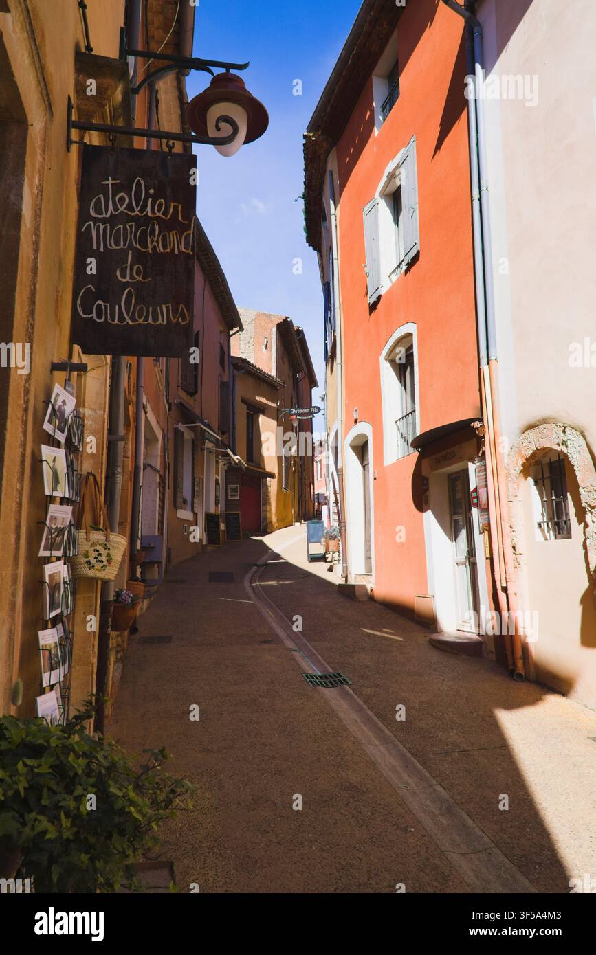Incantevole vicolo stretto con edifici dalle facciate ocra colorate e insegne di negozi nel villaggio collinare di Roussillon, Provenza, Francia. Foto Stock