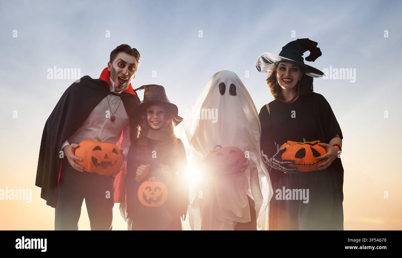 Famiglia gioiosa vestita con costumi creativi di Halloween tra cui un vampiro, streghe e un fantasma, che tiene lanterne di zucca intagliate sorridendo insieme Foto Stock