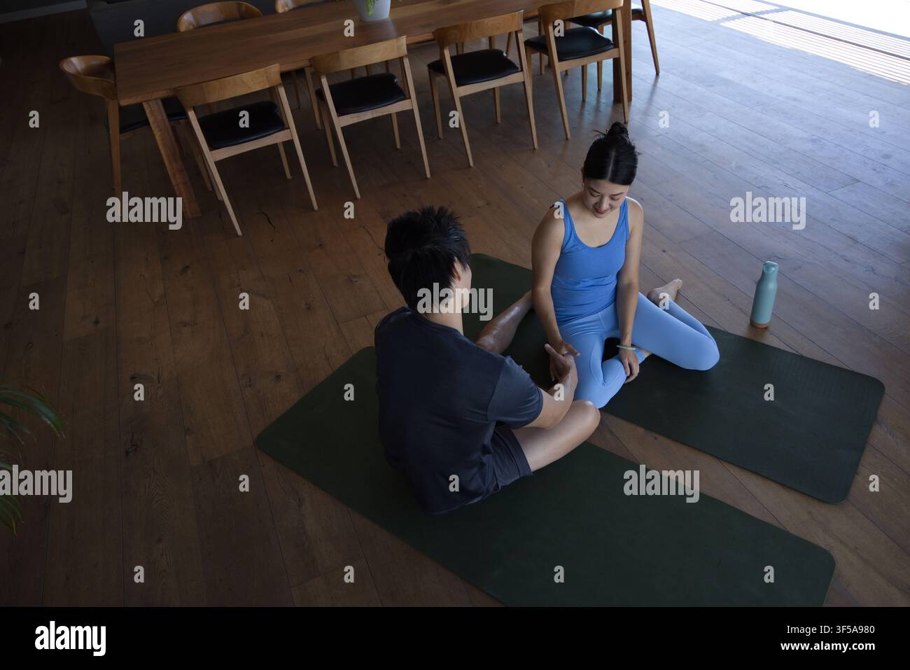Coppia diversa che pratica yoga su tappetini verde scuro a casa vicino al tavolo da pranzo, bottiglia d'acqua Foto Stock
