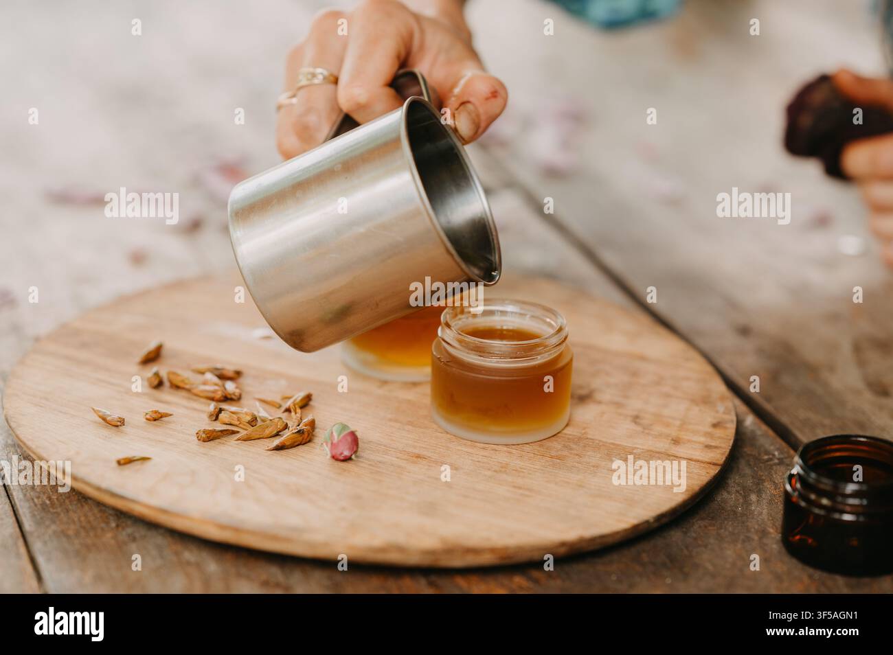 Mano che versa un liquido ambrato in un piccolo barattolo di vetro su una tavola di legno con petali essiccati, mostrando una preparazione cosmetica fatta in casa Foto Stock