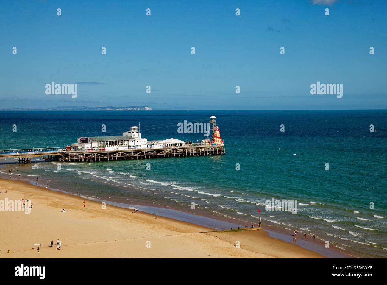 Un gruppo di persone si gode una spiaggia di sabbia dorata con un molo che si protende in un mare blu profondo e scogliere bianche lontane, Bournemouth, Dorset, Inghilterra. Foto Stock