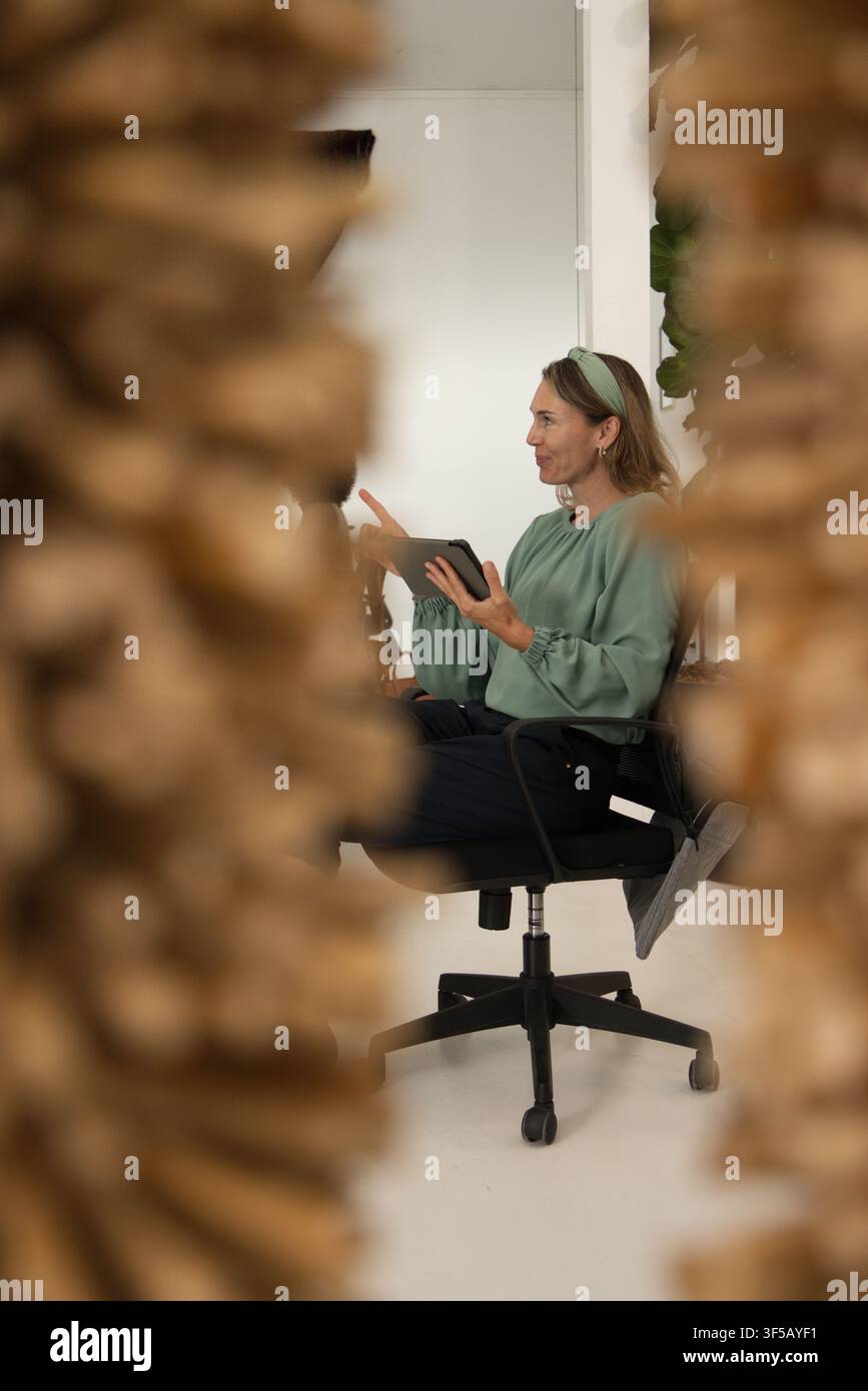 Donna seduta su una sedia girevole nera che tiene un tablet, gesticolando in una stanza luminosa con steli essiccati Foto Stock