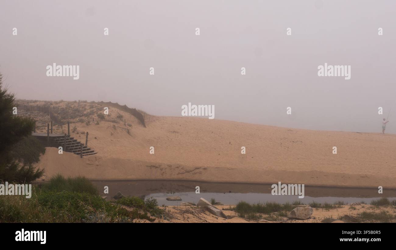 Pescatore solitario che pesca accanto alle dune di sabbia, una scala di legno sepolta e un flusso di marea nella fitta nebbia marina, costa atlantica, Portogallo — Solitudine Foto Stock