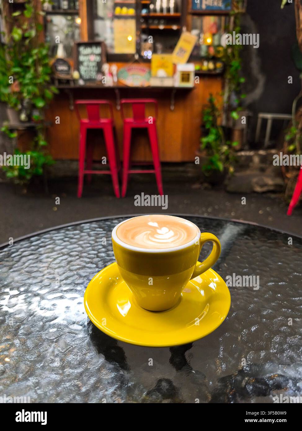 Un latte sapientemente preparato servito in una tazza gialla vivace su un tavolo di vetro, con arte latte cremosa e uno sfondo sfocato di un accogliente caffè rustico Foto Stock