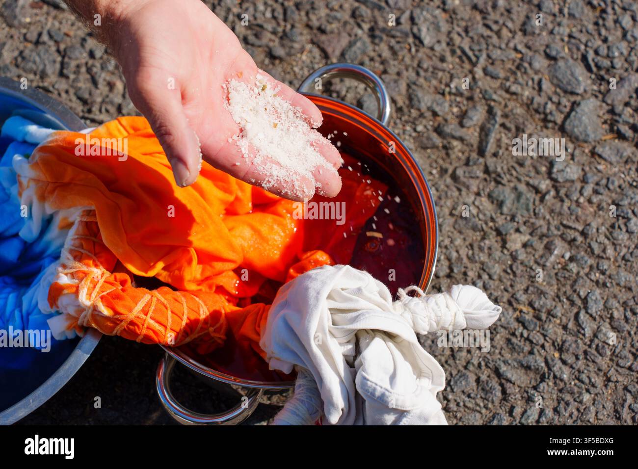 Prima di una mano che aggiunge sale a una pentola contenente un tessuto tie-dye arancione e una tintura rossa, mostrando il processo di fissazione della tintura durante un progetto di fai-da-te Foto Stock