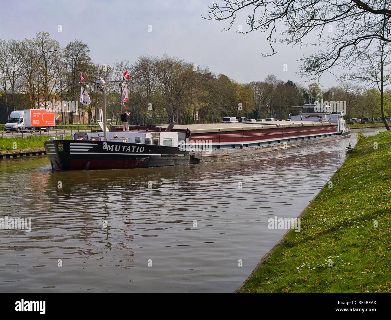 Brugge, Belgio - 09 apr 2026: Una grande chiatta merci naviga lungo un canale fiancheggiato da alberi a Brugge, in Belgio, in una luminosa giornata di primavera Foto Stock