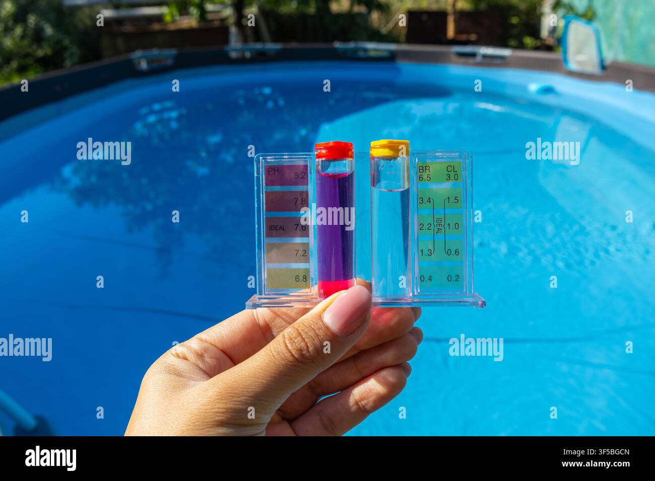 Un primo piano orizzontale di una mano di una persona che tiene un kit di test per il pH e il cloro dell'acqua in plastica trasparente riempito con reagenti liquidi colorati sopra clea Foto Stock