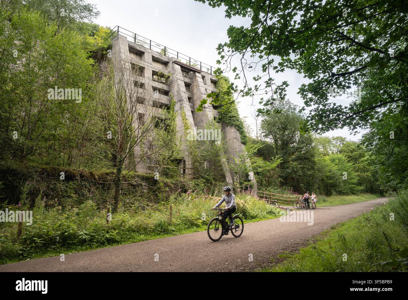 Un ciclista percorre il sentiero di ghiaia del Monsal Trail oltre le imponenti rovine in cemento delle Old East Buxton Lime Kilns, nel Peak District Foto Stock