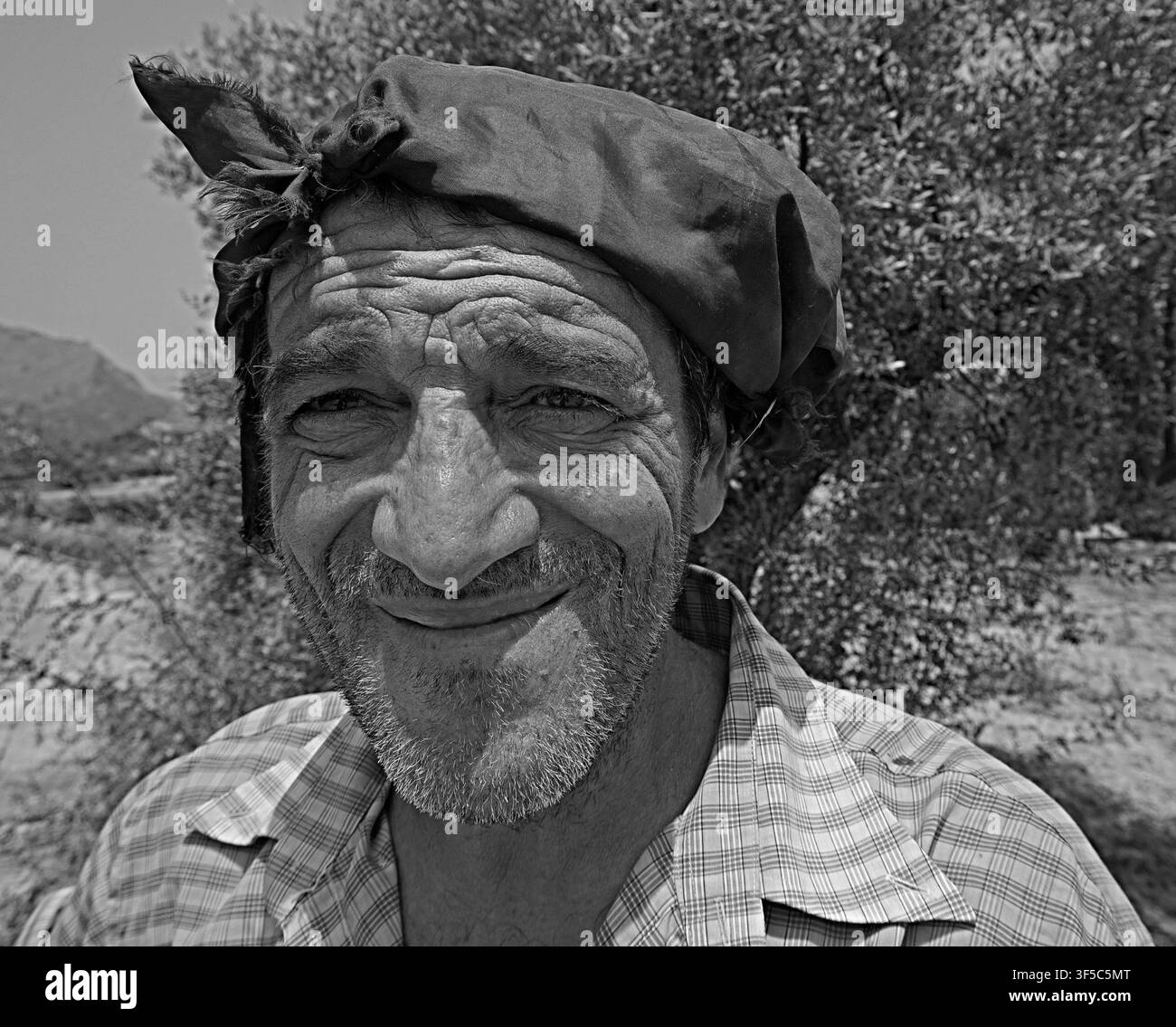 Un ritratto autentico a figura intera di un agricoltore anziano. Il volto del lavoratore è rugoso, sorride calorosamente, indossa jeans strappati e un foulard, e sta Foto Stock