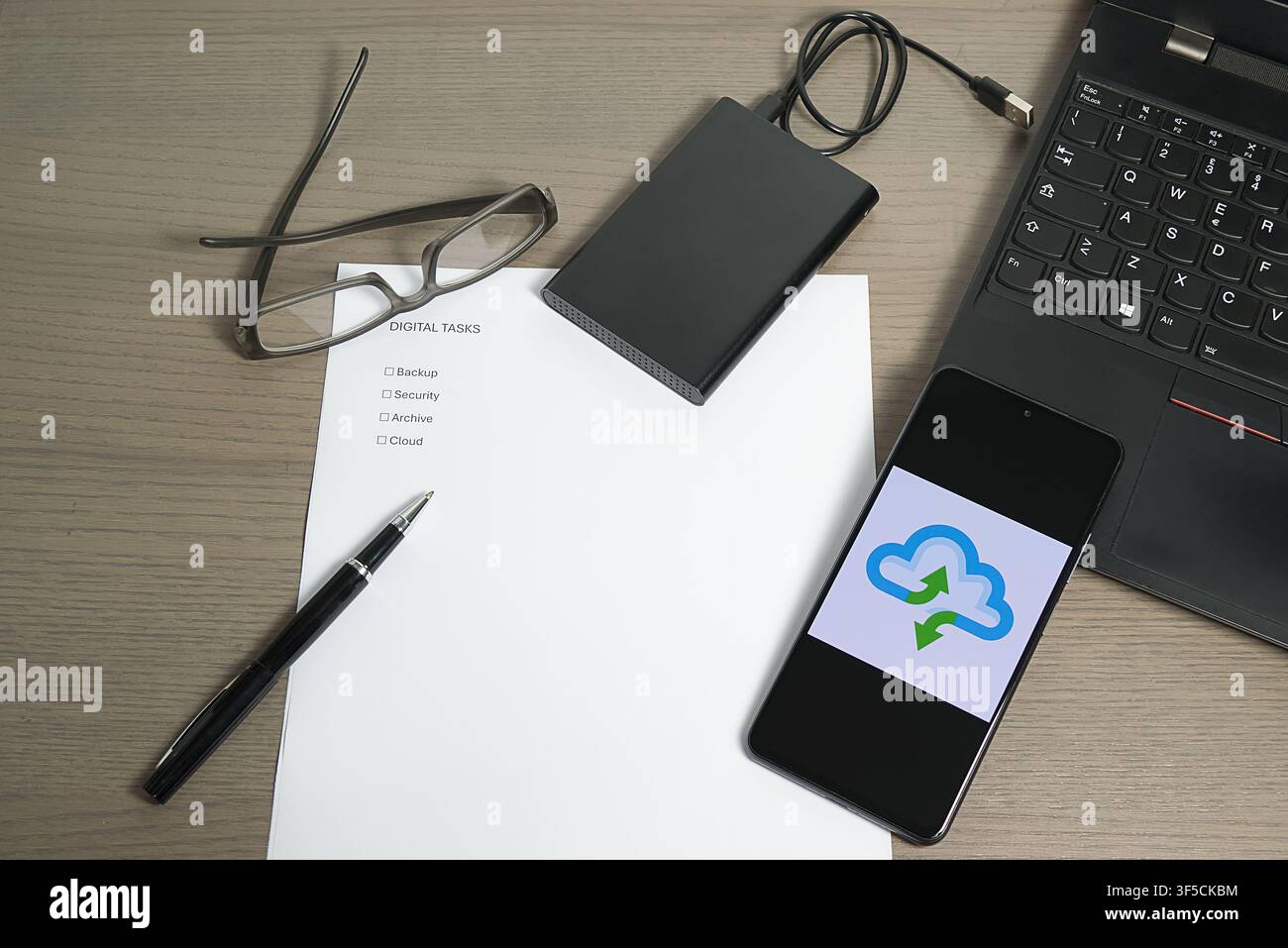 Vista della scrivania con uno smartphone che mostra un'icona a forma di nuvola, una checklist di "Attività digitali" e un disco rigido esterno. Foto Stock