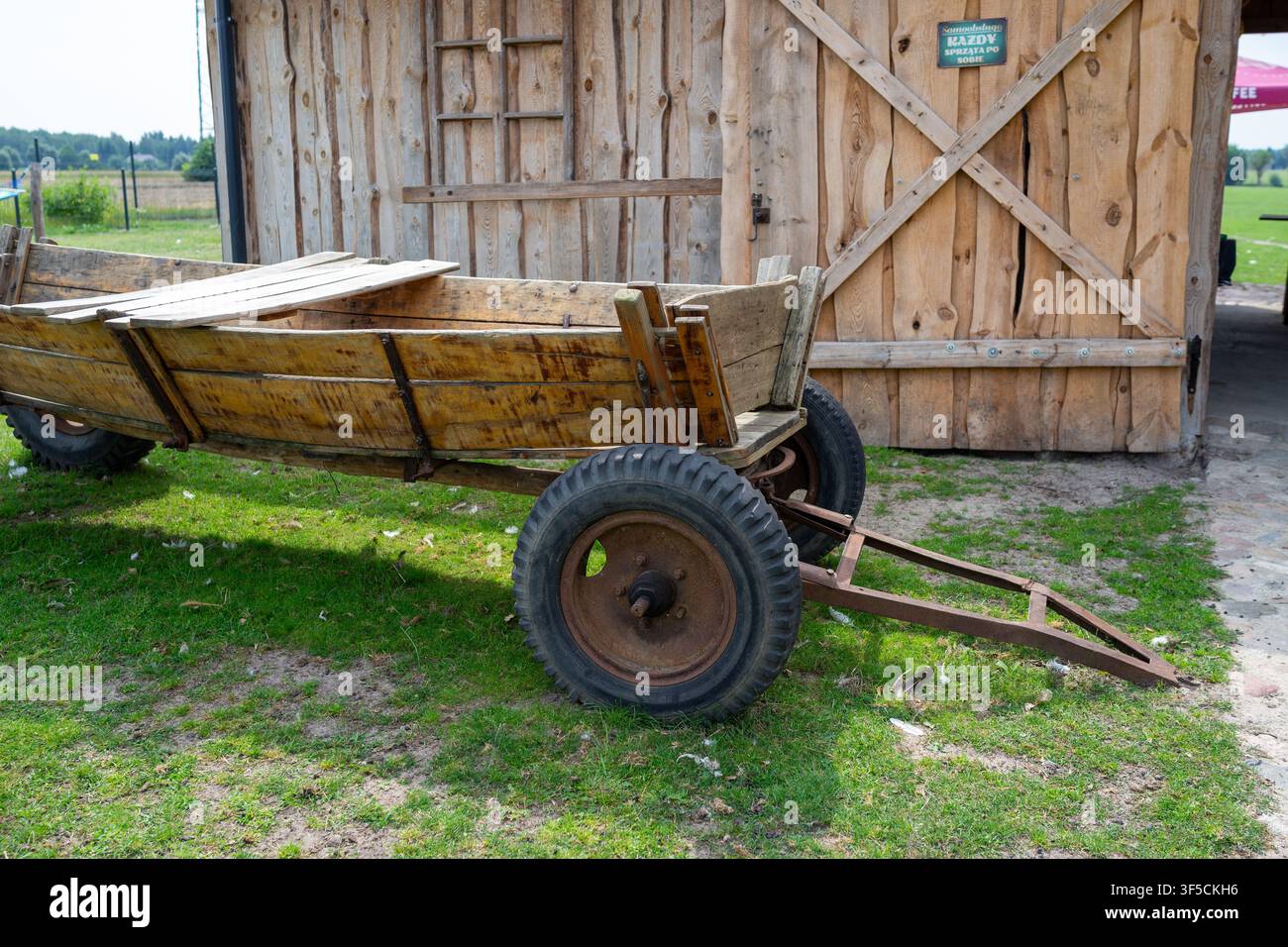 Carro di legno consumato riposa accanto alla porta del fienile. Ruote arrugginite e telaio metallico mostrano un uso intensivo. L'erba verde circonda il carro in un campo aperto. Porta del fienile fe Foto Stock