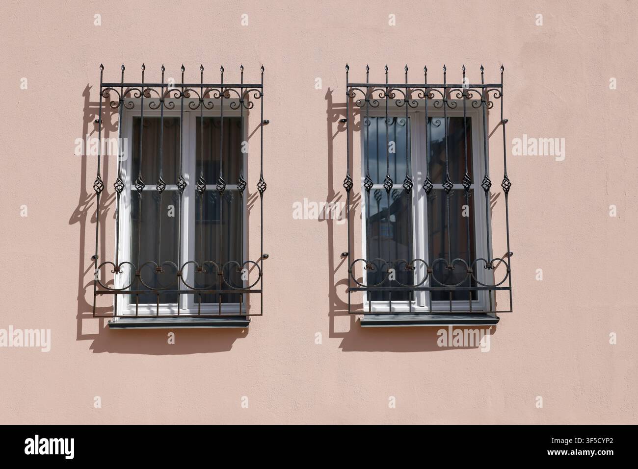 Due finestre sbarre in ferro battuto muro rosa. Griglie di sicurezza vintage in metallo con volute decorative che proiettano ombre nette sulla facciata simmetrica dell'edificio Foto Stock