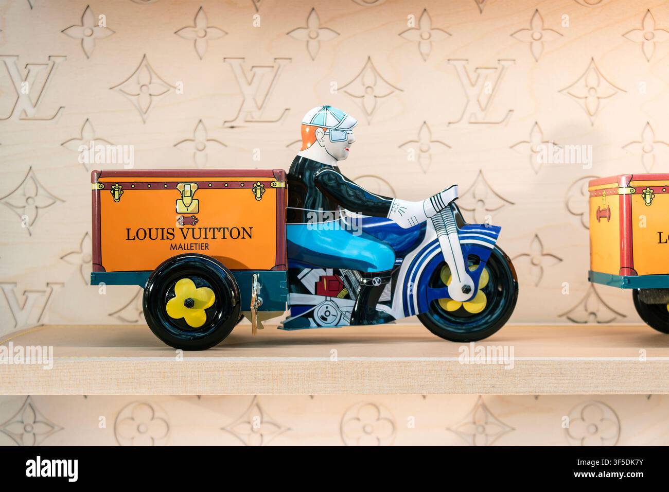 Motocicletta giocattolo vintage in banda di Louis Vuitton con baule di consegna su scaffale di legno che mostra uno sfondo con monogramma Foto Stock