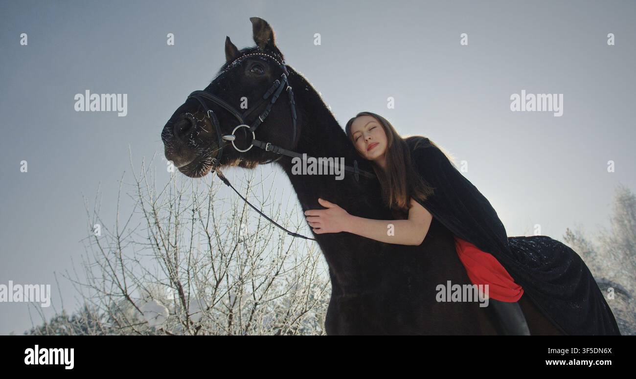 Donna che abbraccia un cavallo nero in una tranquilla foresta invernale Foto Stock