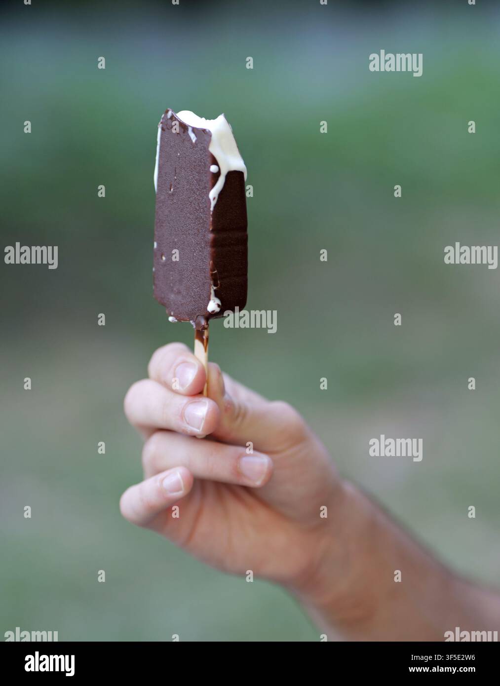 Mano che tiene una deliziosa barretta di gelato alla vaniglia ricoperta di cioccolato parzialmente mangiata su un bastoncino Foto Stock