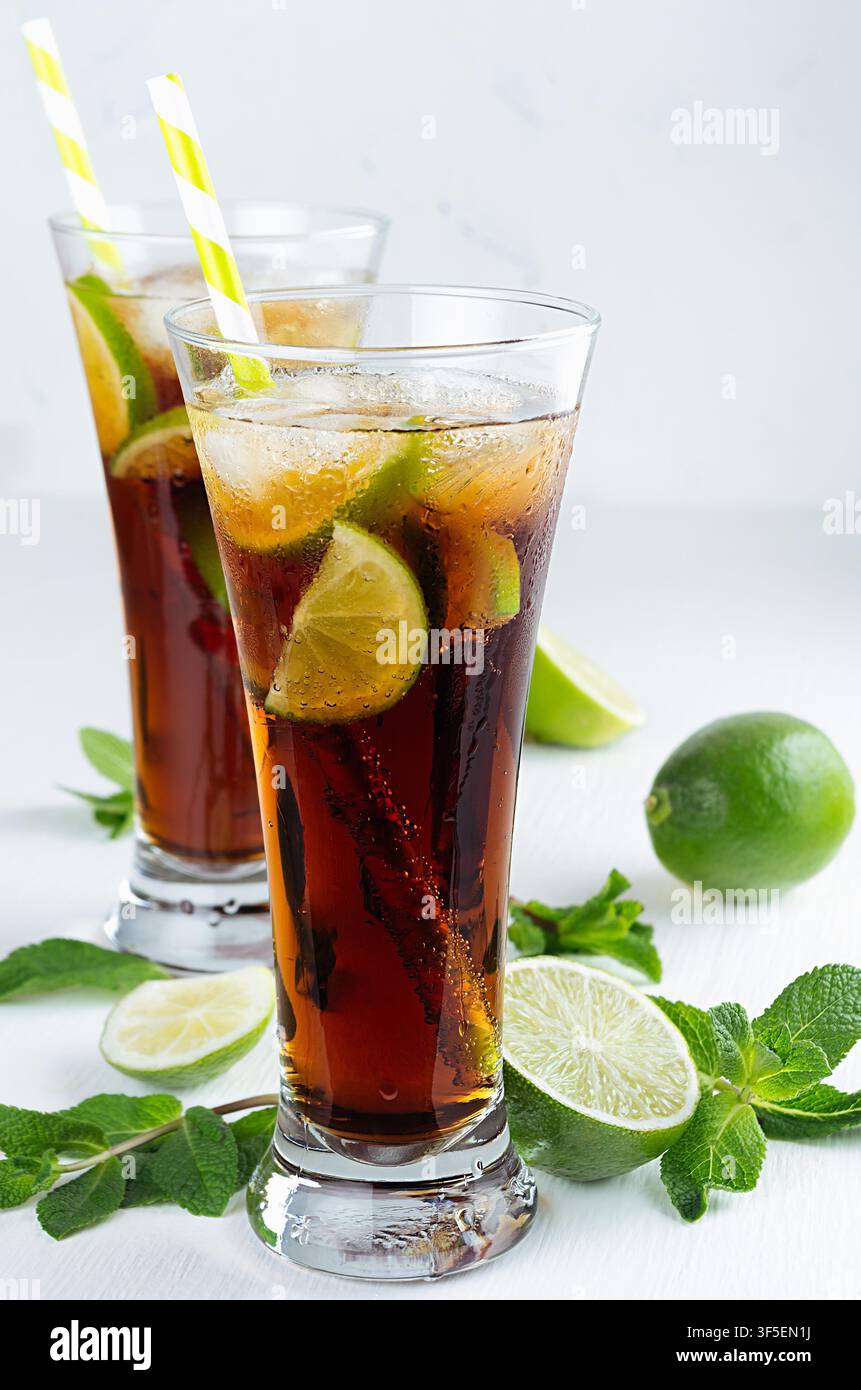 Cocktail freddo cuba libre in due lunghi bicchieri bagnati con paglia, pezzi di lime, menta su morbido sfondo bianco moderno. Foto Stock