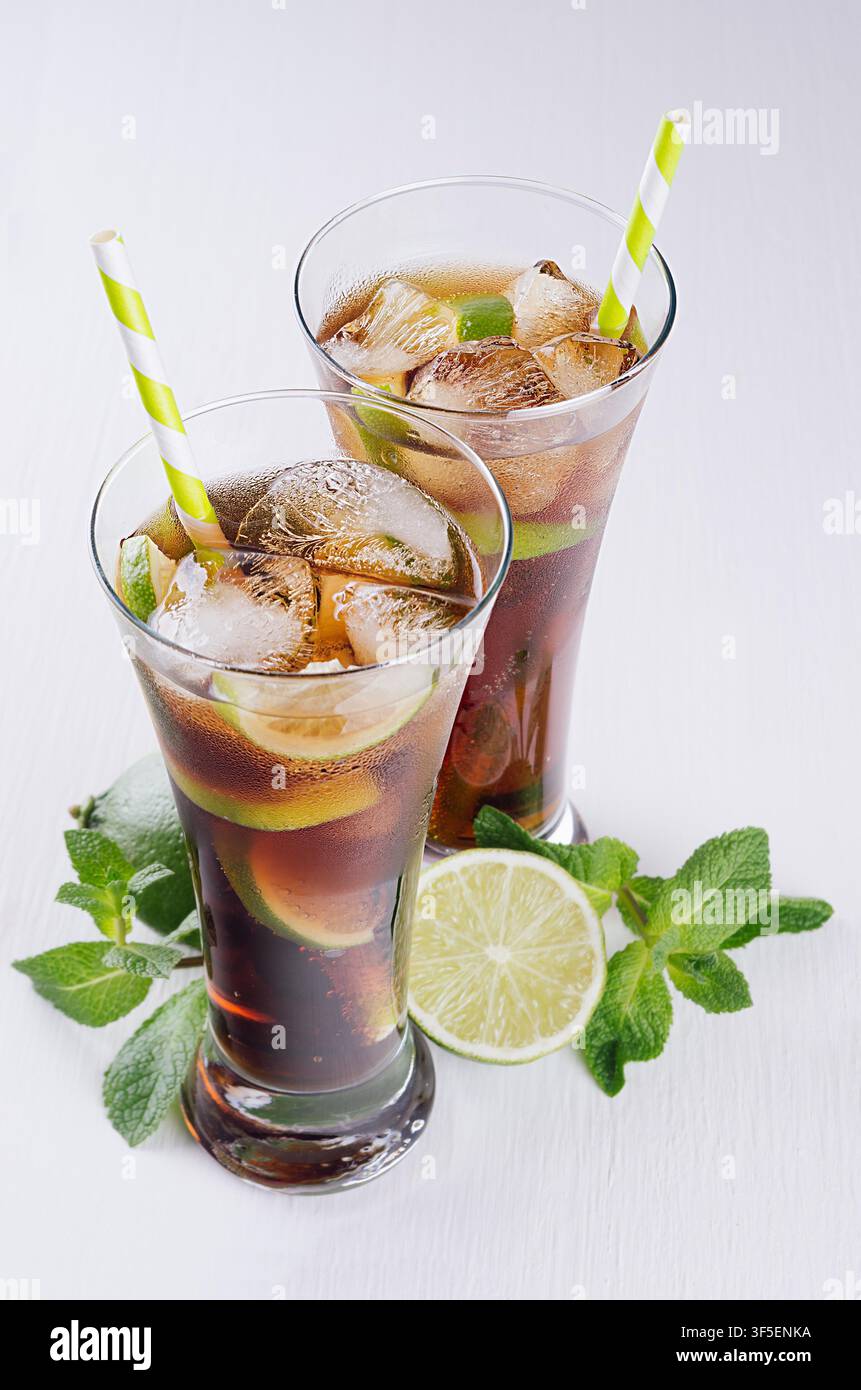 Cocktail freddo cuba libre in due bicchieri bagnati con paglia, pezzi di lime, menta su morbido sfondo bianco moderno, vista dall'alto, primo piano. Foto Stock