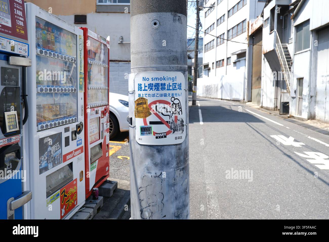Un palo dell'utenza con un cartello "Divieto di fumare" con testo giapponese, situato accanto a distributori automatici giapponesi lungo una tranquilla strada laterale di Tokyo. Foto Stock