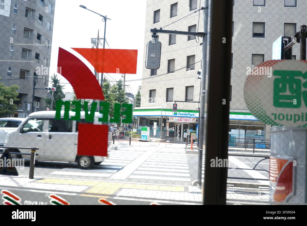 Un furgone bianco che passa davanti a un angolo di strada con un FamilyMart visibile dall'altra parte dell'incrocio, scattato attraverso il logo della vetrina di un negozio 7-Eleven in Foto Stock