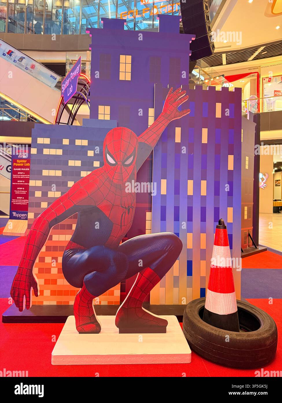 Un cartone ritagliato di Spider-Man posato in posizione accovacciata davanti a uno sfondo paesaggistico cittadino colorato all'interno di un centro commerciale. - Immagine stock catturata con smartphone