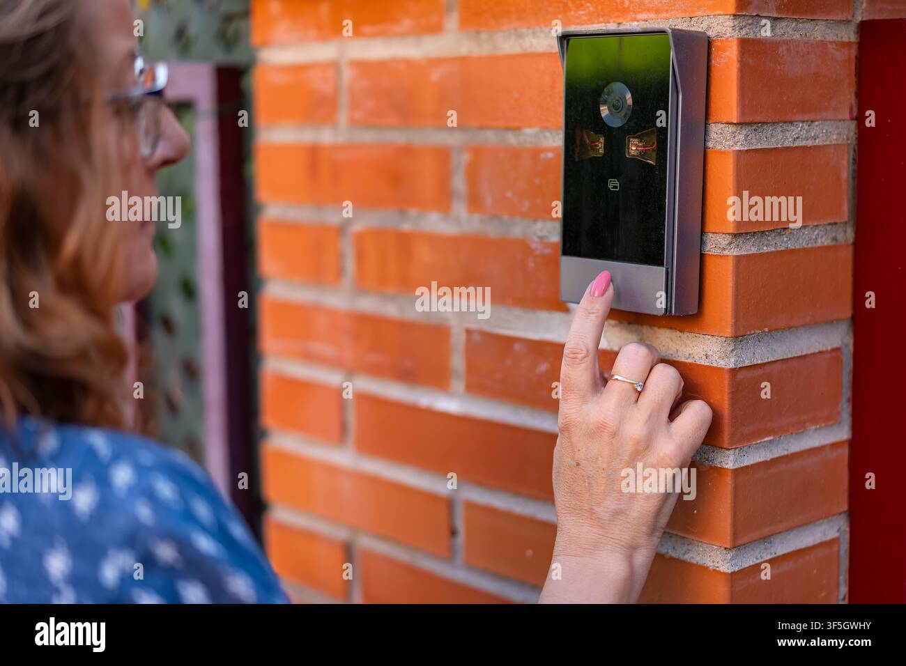 Donna che preme un campanello video moderno montato su un muro d'ingresso in mattoni arancioni all'aperto Foto Stock