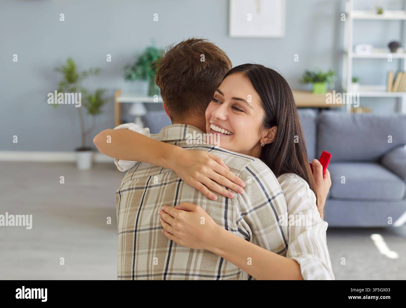 Coppia che si abbraccia dopo la proposta di matrimonio tenendo un anello e un regalo a casa Foto Stock