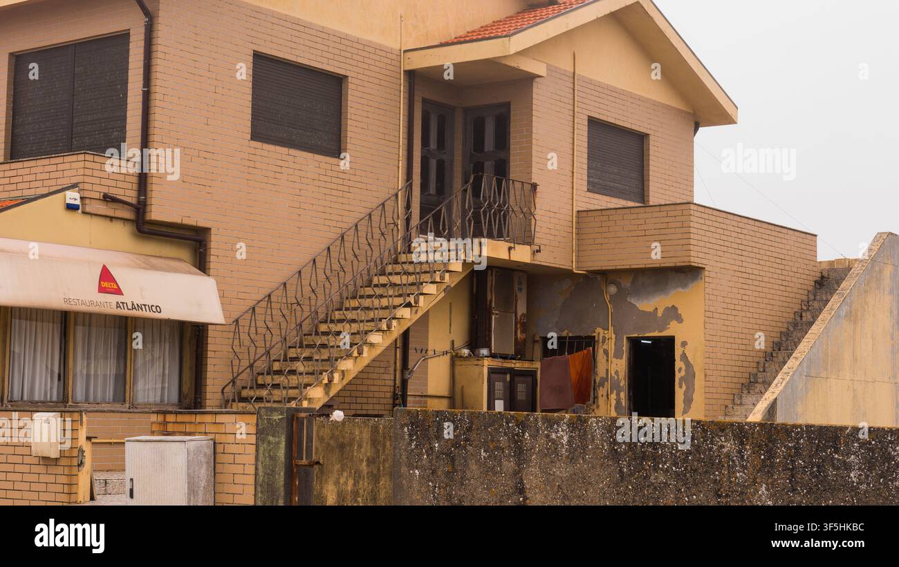 Casa in mattoni marroni con scala esterna, serrande a rullo e intonaco danneggiato dall'umidità sopra un piccolo ristorante, Nord Portogallo Foto Stock