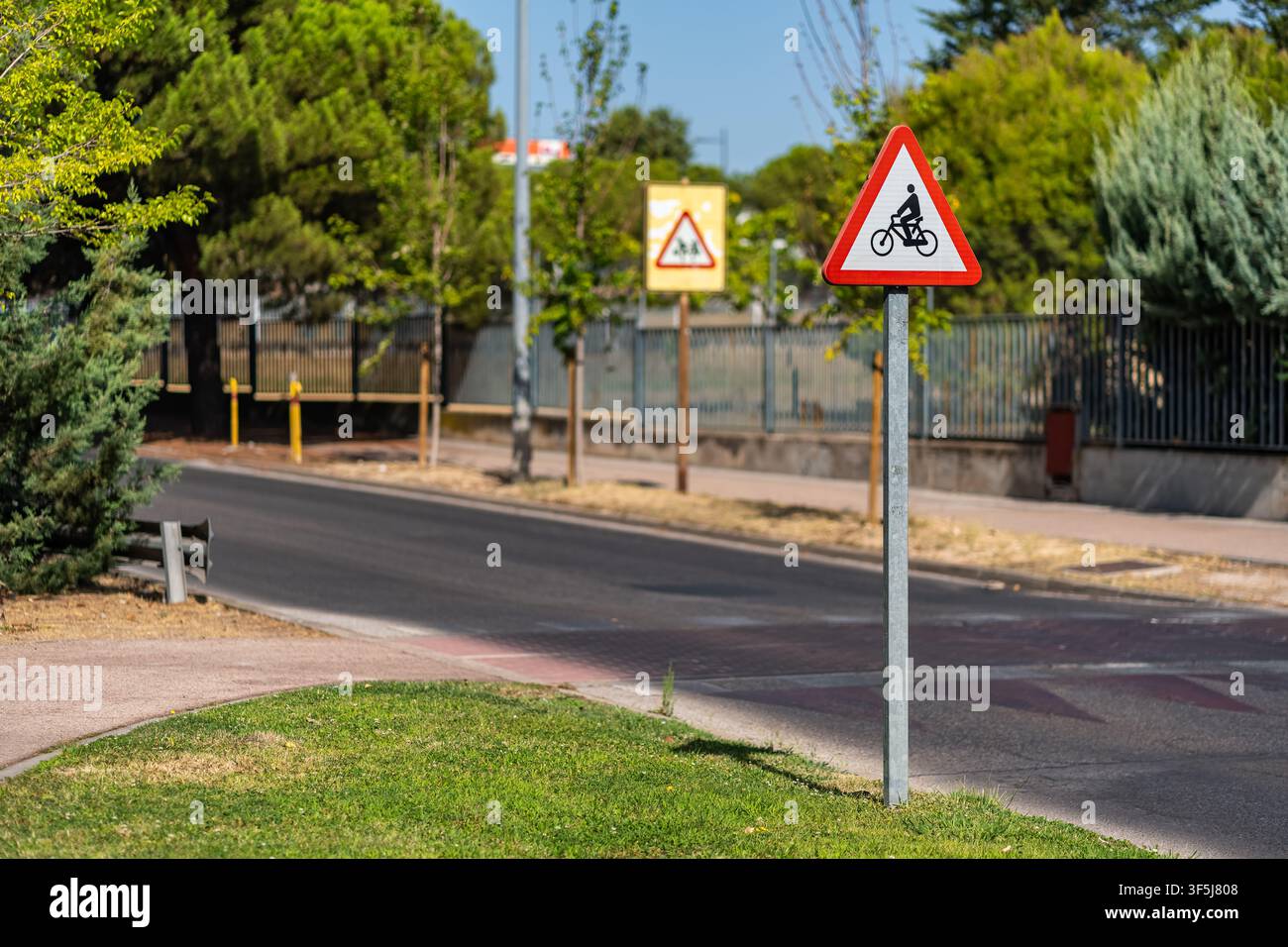 Segnale di avvertimento per biciclette accanto a una strada urbana e attraversamento ciclabile segnalato circondato da alberi Foto Stock