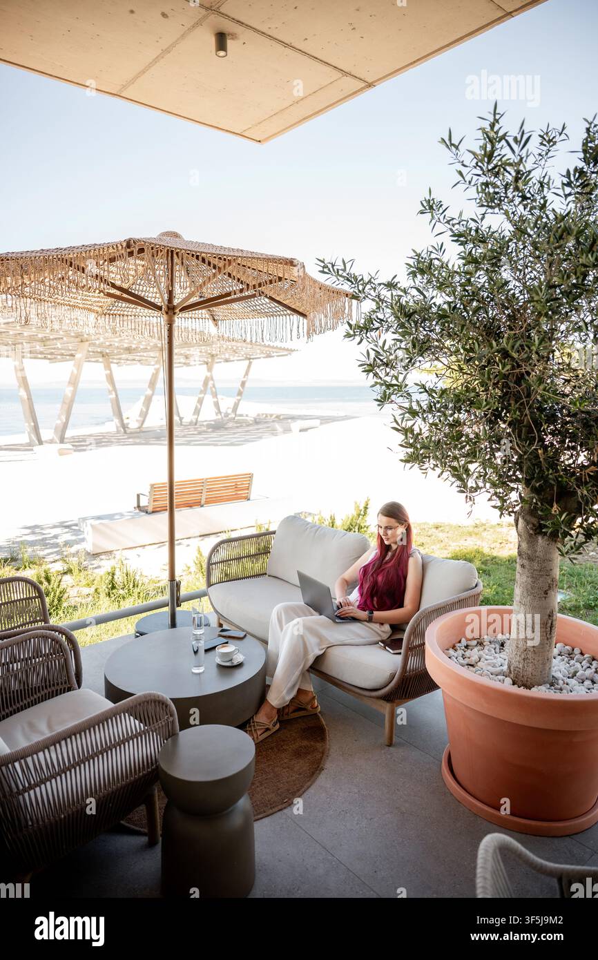 Una freelance si rilassa in un chic caffè sul mare, incarnando l'equilibrio della vita e lo stile vecchio denaro. Lavora sul suo laptop mentre gode dell'aria serena costiera Foto Stock