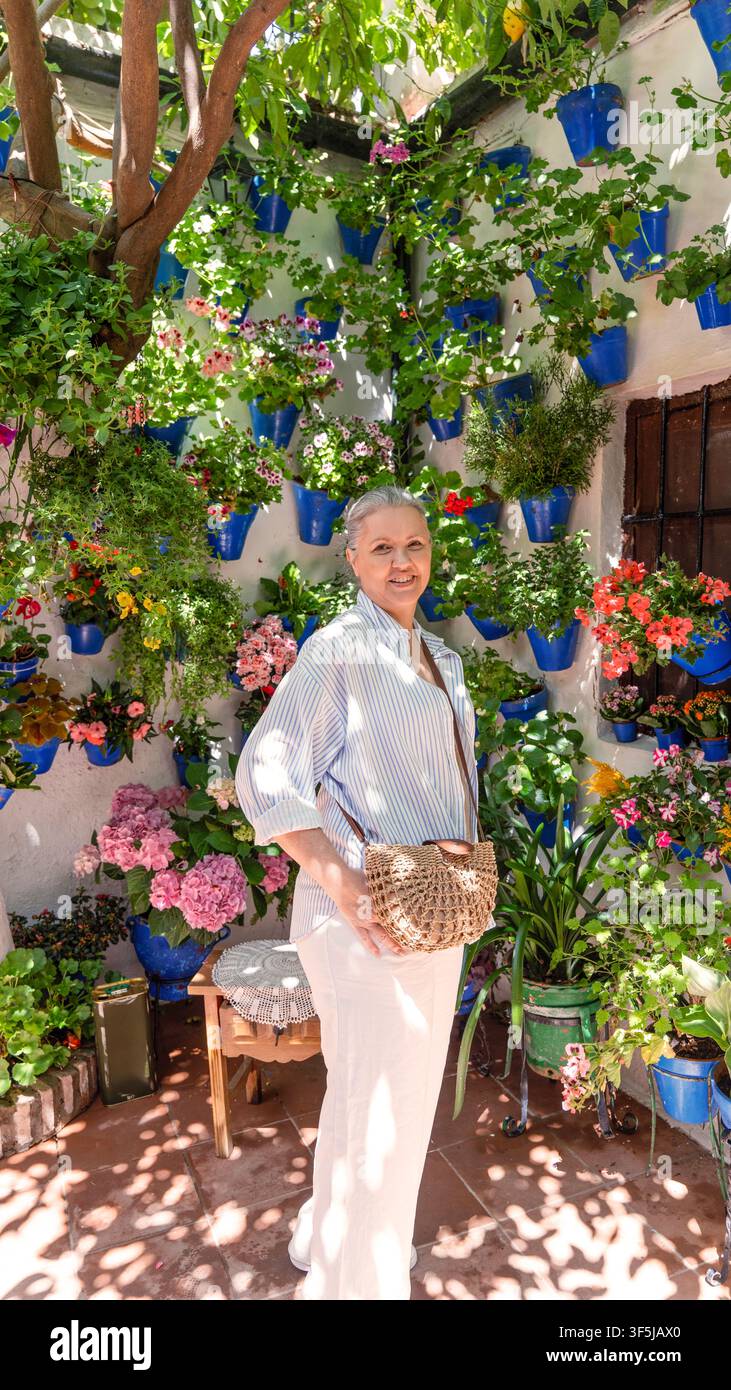 Donna anziana che tiene un cesto di vimini in un cortile illuminato dal sole a Córdoba, circondata da fiori colorati, vegetazione lussureggiante, mura bianche e iconiche fiori blu Foto Stock