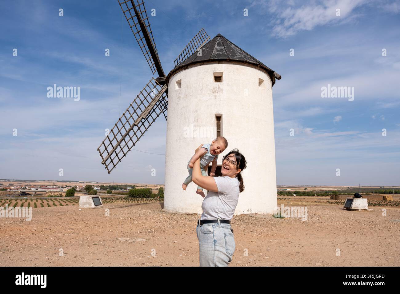 Madre felice che cresce un bambino davanti a un mulino a vento tradizionale spagnolo sotto un cielo blu Foto Stock
