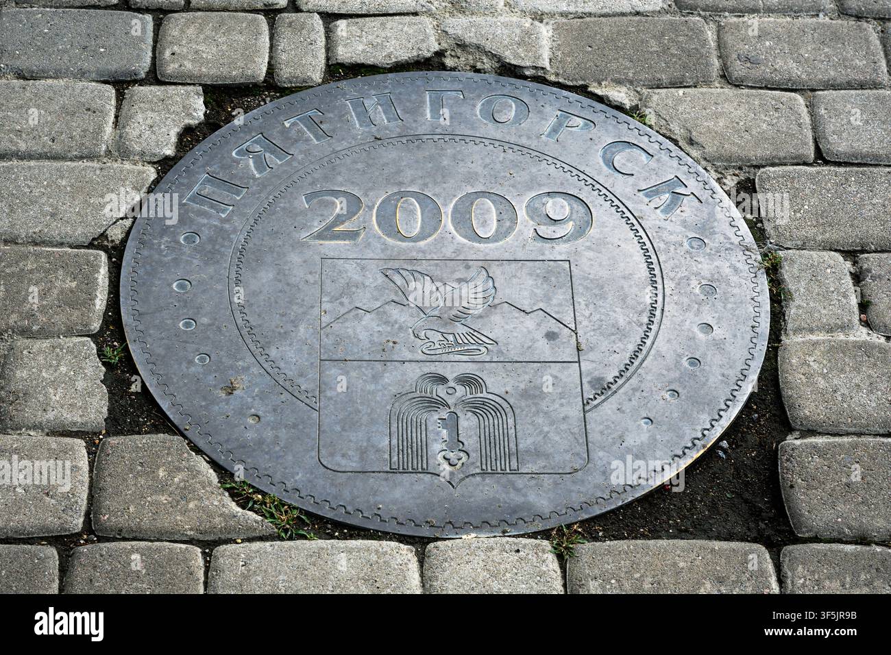 Una vista ravvicinata dall'alto di una placca metallica circolare incastonata in un marciapiede di ciottoli grigi. Il disco metallico consumato presenta il testo cirillico "ПЯТ"} Foto Stock