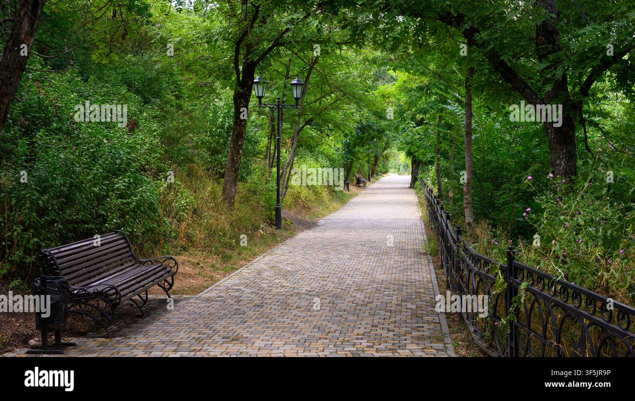 Un marciapiede lastricato si snoda attraverso un parco rigoglioso e verde, fiancheggiato da alberi e cespugli fitti che creano una chioma naturale. Il sentiero è costruito fr Foto Stock