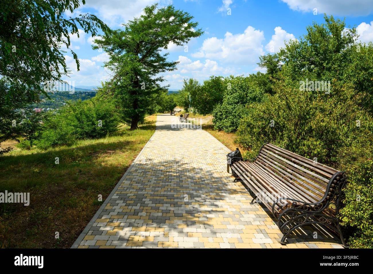 Una fotografia luminosa e soleggiata di un marciapiede lastricato vuoto in un parco pubblico. Una panchina di legno con una struttura metallica si trova sul lato destro del sentiero, accanto a un Foto Stock
