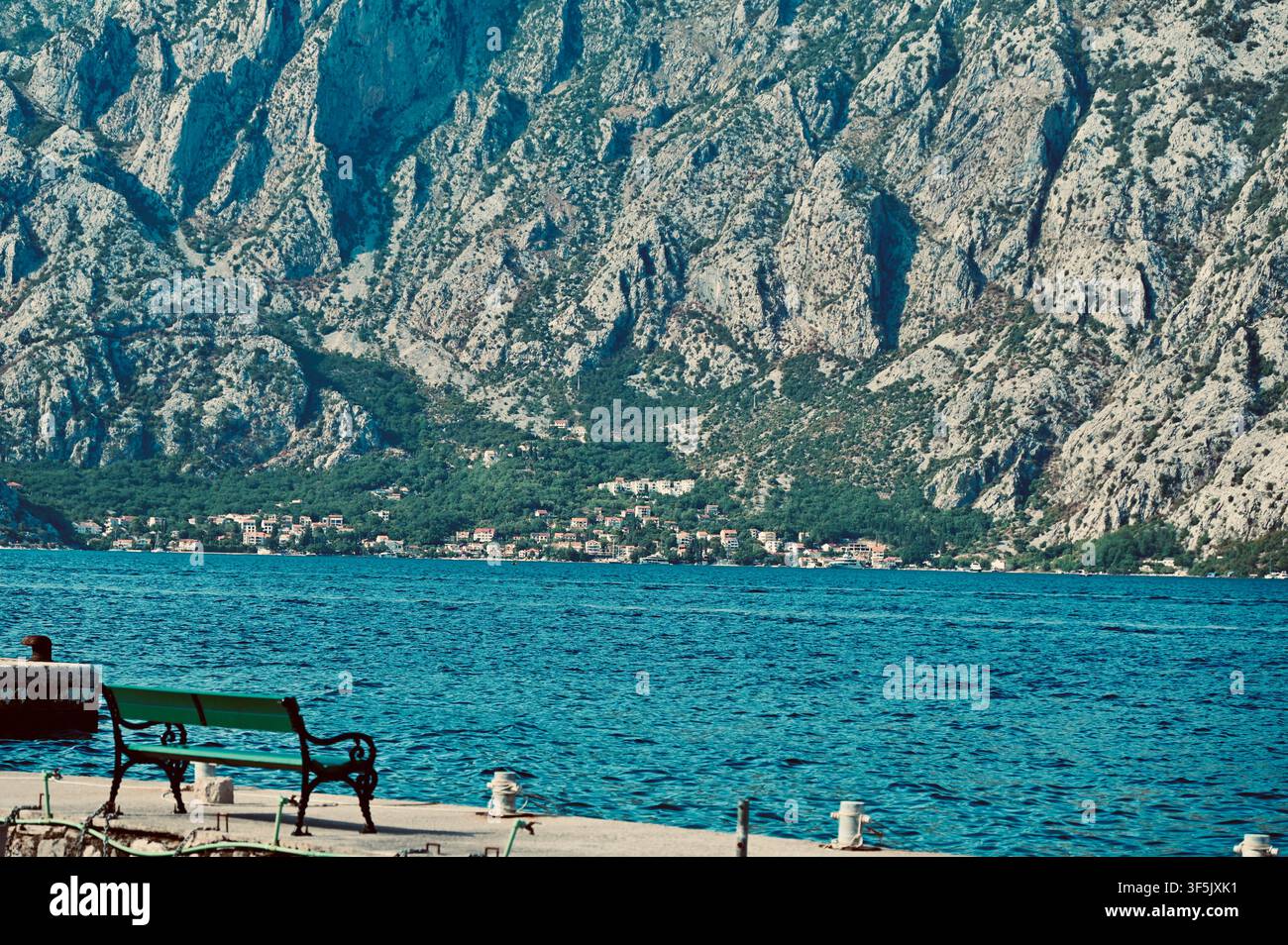 Un ampio paesaggio orizzontale con una panchina verde vuota in primo piano su un molo di pietra, che si affaccia su un mare blu brillante con una città costiera e imponenti montagne rocciose sullo sfondo Foto Stock