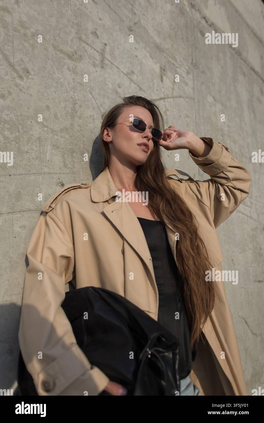 Scatto medio di una giovane donna elegante con lunghi capelli che indossa un trench beige e occhiali da sole, in posa contro un muro di cemento sotto la luce intensa del sole. Urb Foto Stock