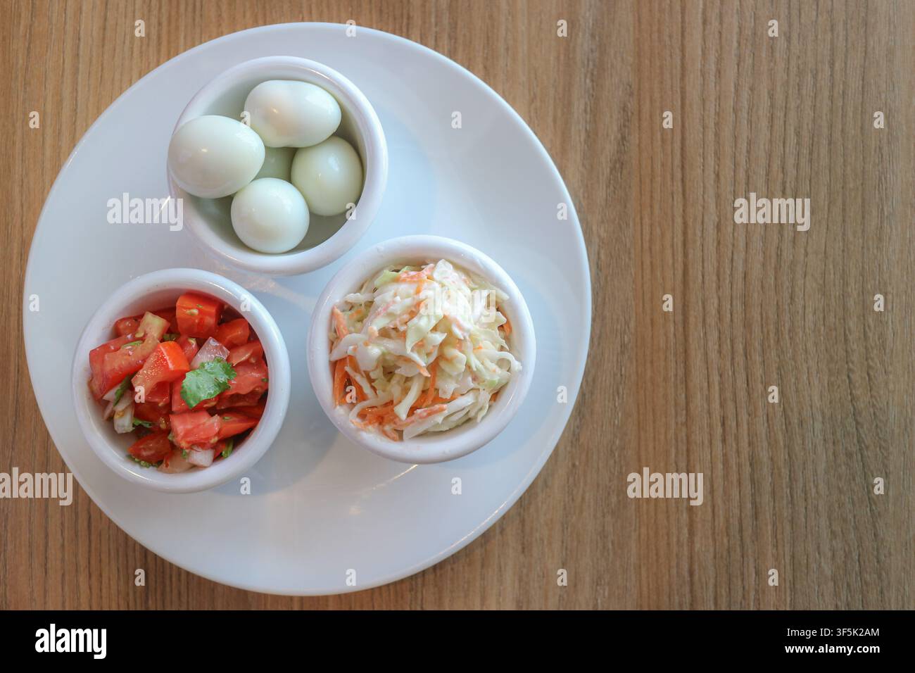 Vista dall'alto di salsa al pomodoro, uova di quaglia bollite e insalata di cavolo in ciotole separate su un piatto di ceramica bianca su un tavolo di legno chiaro, che mostra cibo fresco. Foto Stock